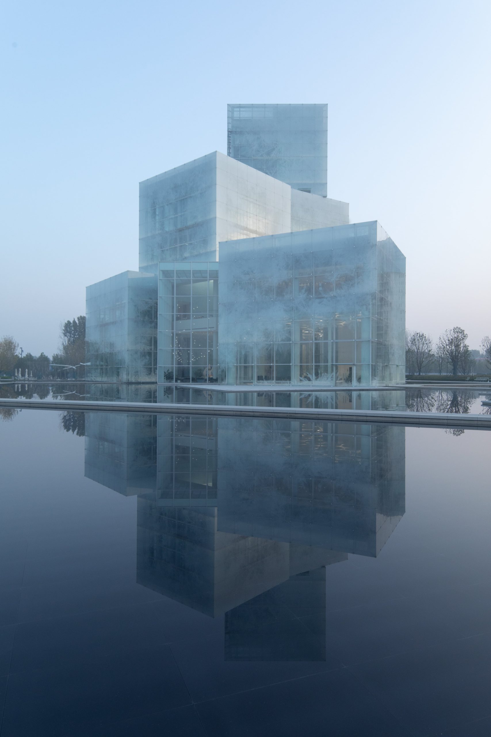 Blick auf ein aus einzelnen Würfeln zusammengesetztes Glasgebäude vor einer Waserfläche, in der es sich spiegelt. Mathieu Forest Architectes und Zone Of Utopia, Xinxiang Cultural Tourism Center, Foto: ArchExist